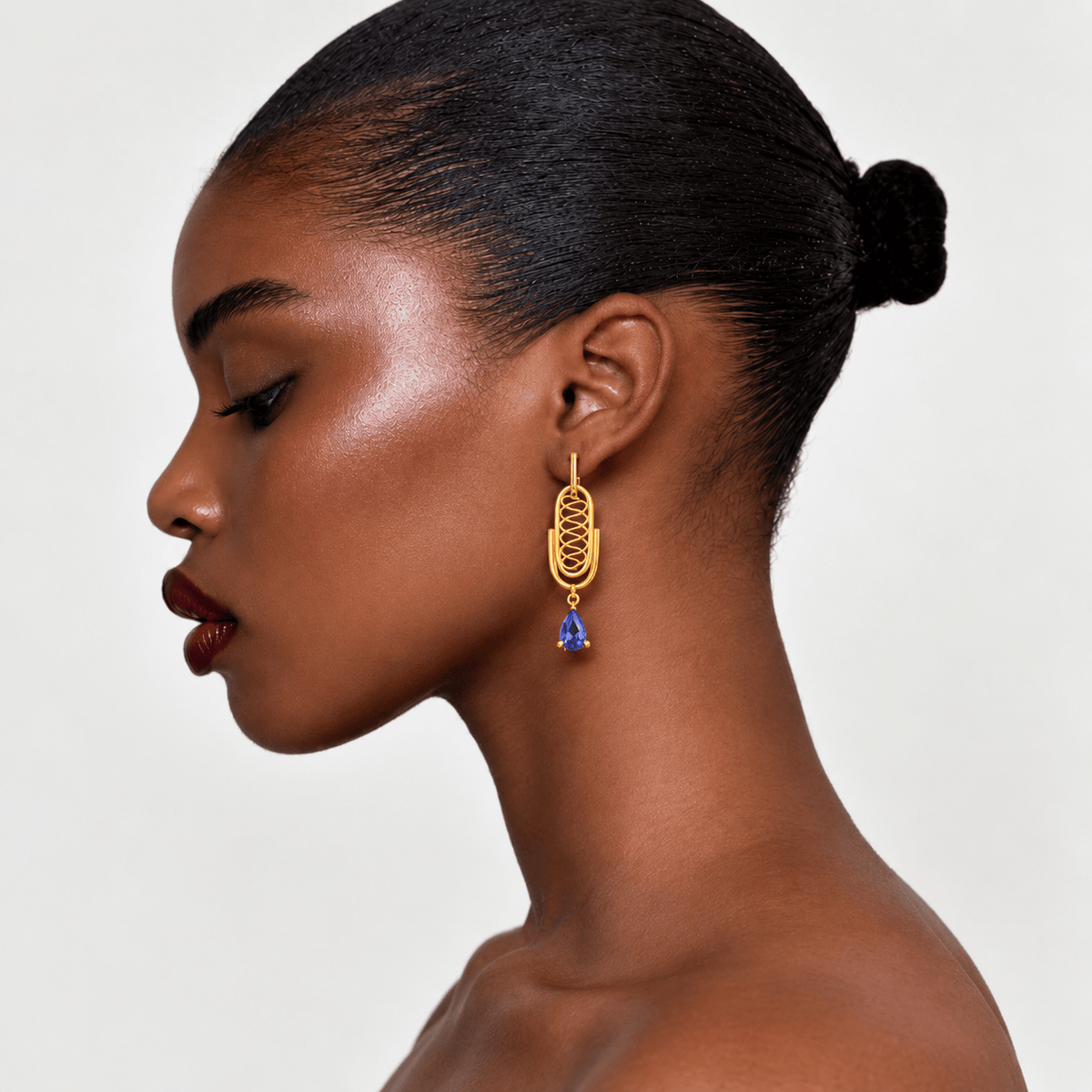 A close-up side profile of a model wearing a gold dangle earring with a blue teardrop gemstone, set against a clean light gray background.