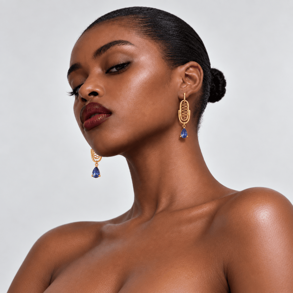A close-up portrait of a woman wearing gold dangle earrings with blue teardrop charms against a light gray background.