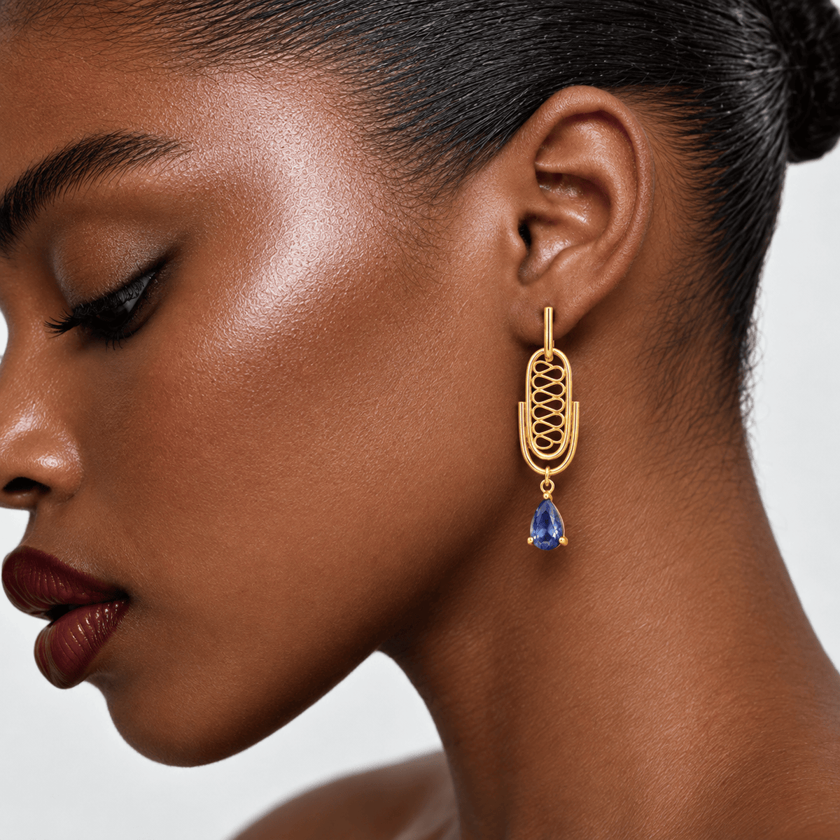 A close-up side profile of a woman wearing a gold dangle earring with a blue teardrop gemstone, against a clean light gray background.