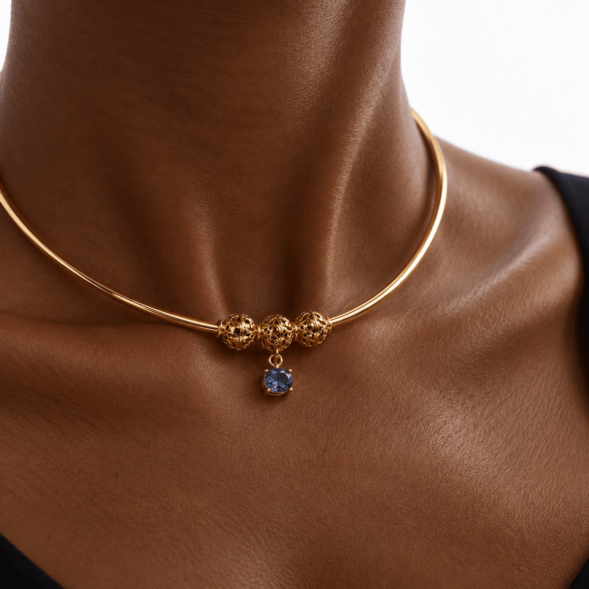 A close-up of a gold bangle necklace with three textured gold beads and a dangling blue gemstone charm, shown on a model's neck.