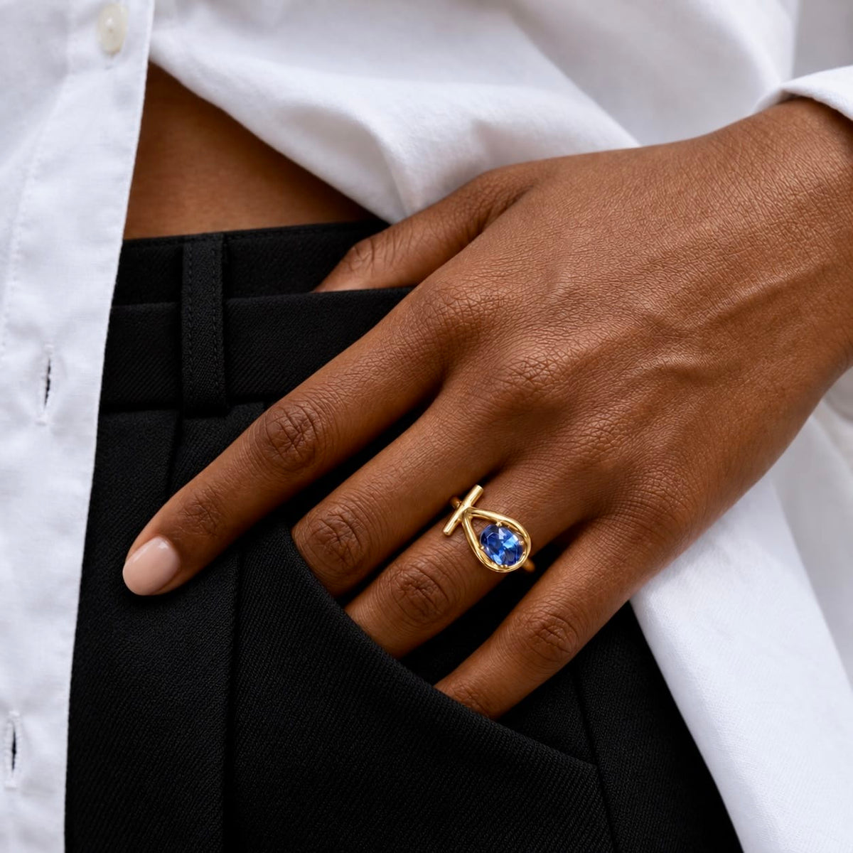 A close-up of a person's hand wearing a gold ring with a teardrop-shaped blue gemstone, resting on their black trousers and white shirt.