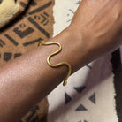 A brass wrist bracelet with a flowing curve design, worn on a person's wrist, against a patterned fabric background.