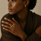 A close-up lifestyle shot of a model wearing a gold ring with a circular band and a diagonal bar, styled with a matching gold hoop earring and a dark brown blazer.