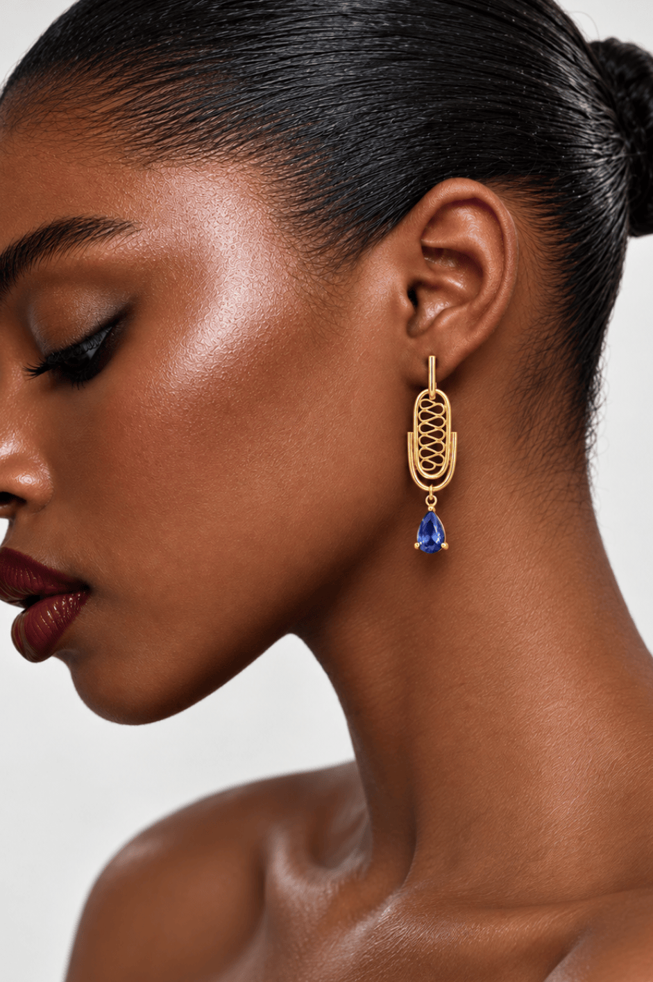 A close-up side profile of a model wearing a gold drop earring with a tanzanite teardrop gemstone, against a clean light gray background.
