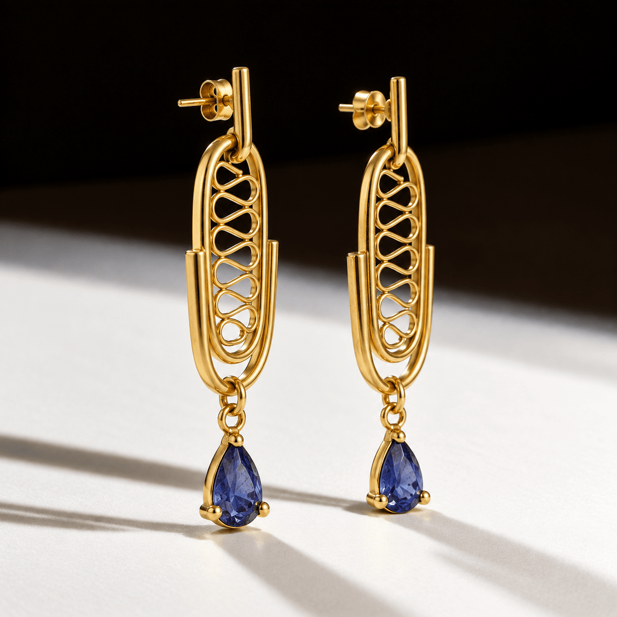 A pair of gold drop earrings with an openwork geometric frame and a teardrop-shaped blue gemstone hanging from each earring, photographed on a white surface with a dark background.