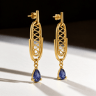 A pair of gold drop earrings with an openwork geometric frame and a teardrop-shaped blue gemstone hanging from each earring, photographed on a white surface with a dark background.