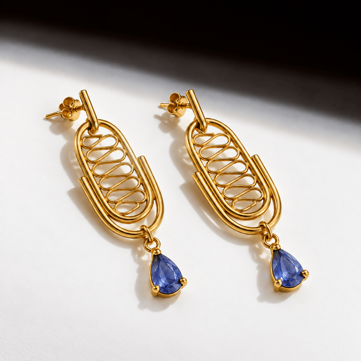 A pair of gold drop earrings with a double-ring oval design and a teardrop-shaped blue gemstone dangling from each earring, photographed on a white surface with a dark shadow in the upper right.