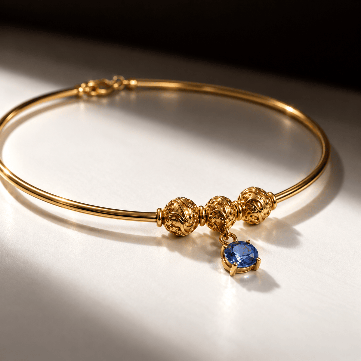 A gold-plated bangle bracelet with three ornate gold beads and a dangling blue gemstone charm, displayed on a white surface with dramatic shadows.