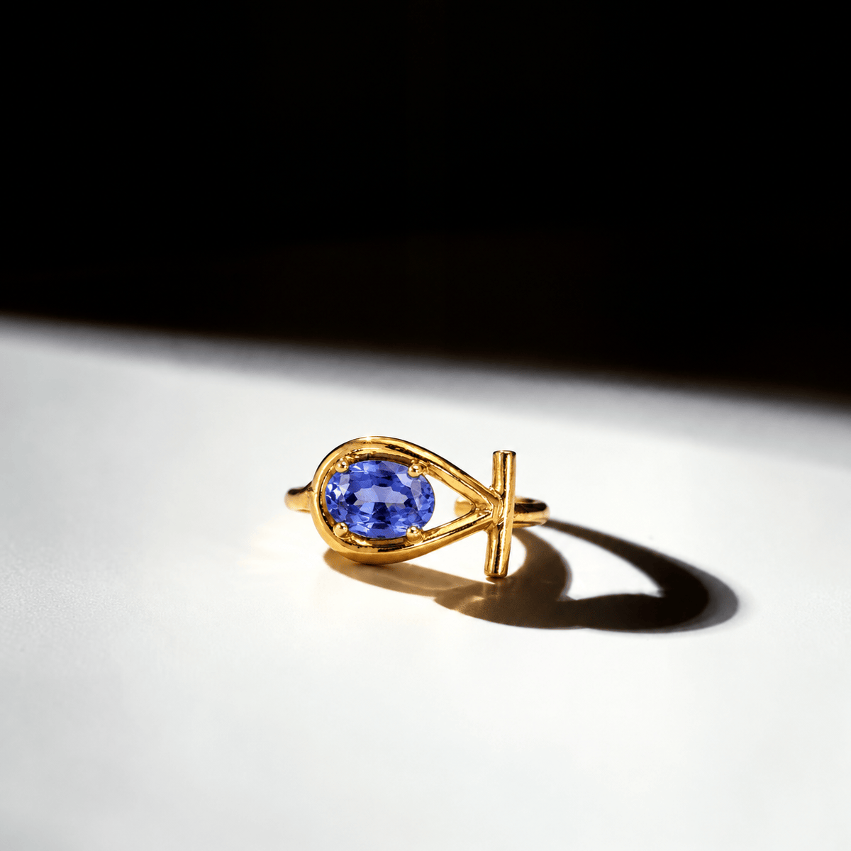 A gold ring featuring a teardrop-shaped setting with a round blue gemstone, photographed on a white surface under dramatic spotlighting with a strong shadow.