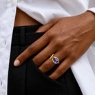 A close-up of a person's hand wearing a gold ring with a teardrop-shaped blue gemstone, resting on their black trousers and white shirt.
