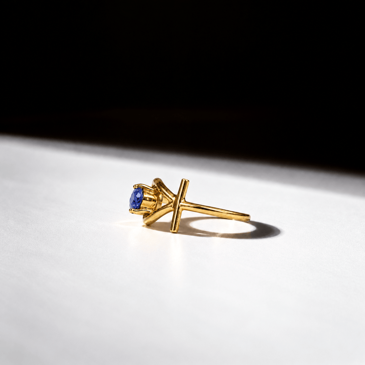 A gold ring with a single blue gemstone in a unique open cross-like setting, photographed on a white surface with dramatic shadow and a black background.