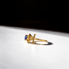 A gold ring with a single blue gemstone in a unique open cross-like setting, photographed on a white surface with dramatic shadow and a black background.