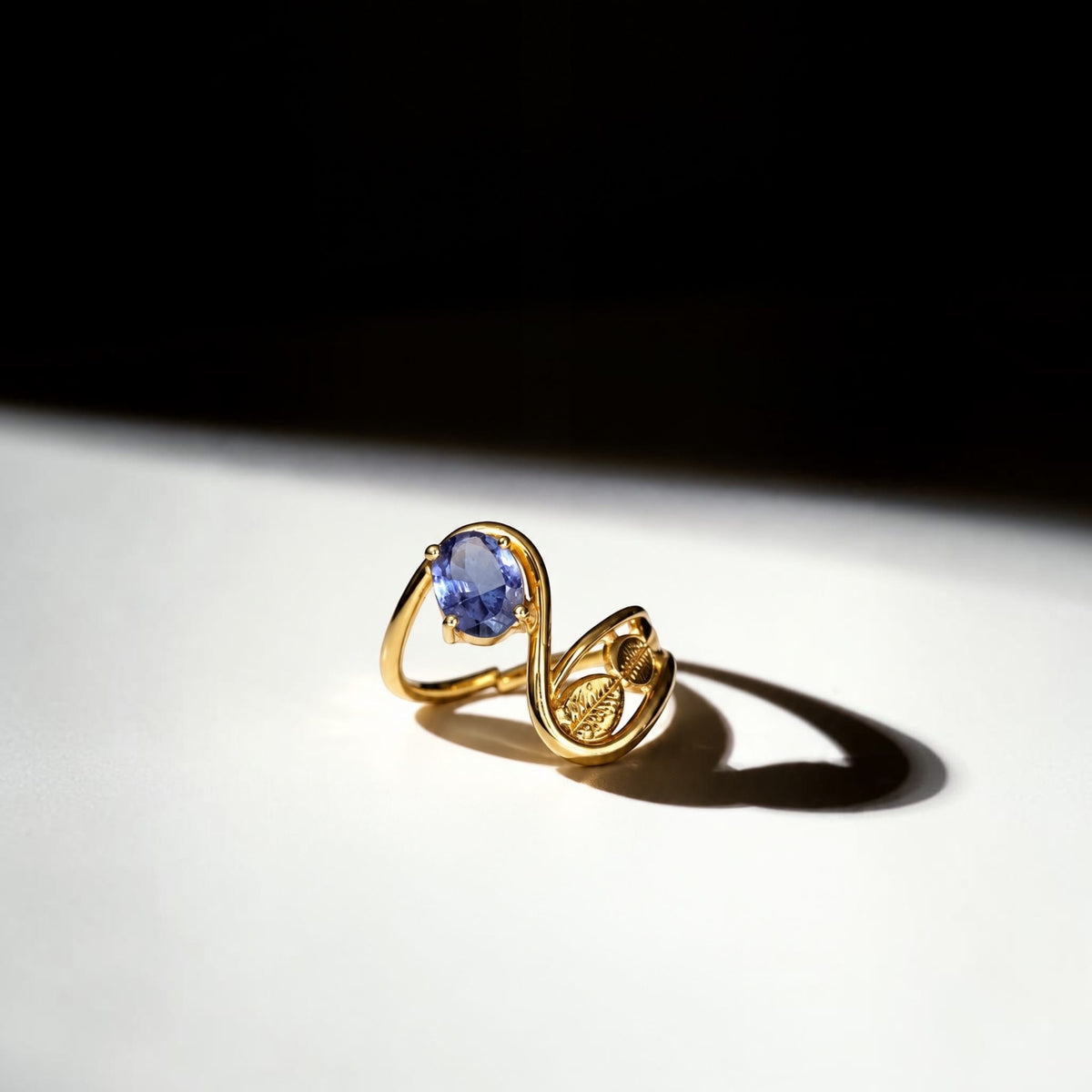 A gold ring with a pear-cut blue gemstone, photographed on a white surface under dramatic side lighting with a strong shadow against a black background.