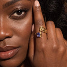 A close-up lifestyle shot of a woman wearing a gold ring with a round blue gemstone on her finger, with her face and hand prominently framed.