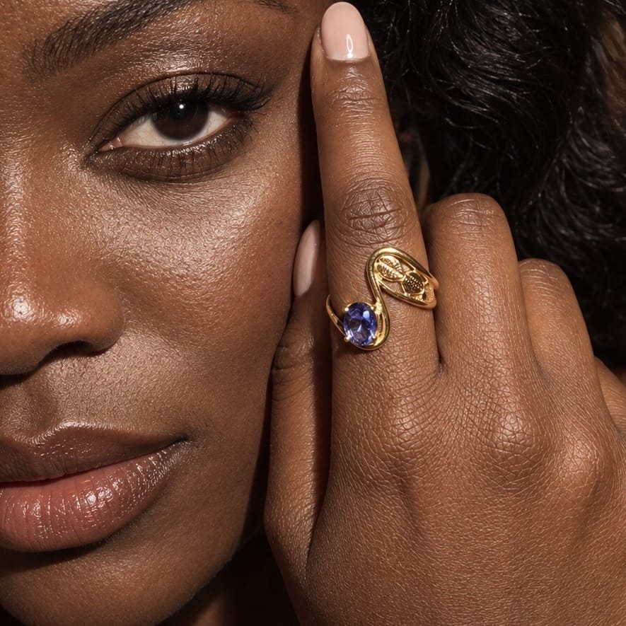 A close-up lifestyle shot of a woman wearing a gold ring with a round blue gemstone on her finger, with her face and hand prominently framed.