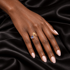 A close-up of a hand with pale pink manicured nails resting on draped black satin fabric, wearing a gold ring with a blue oval gemstone.
